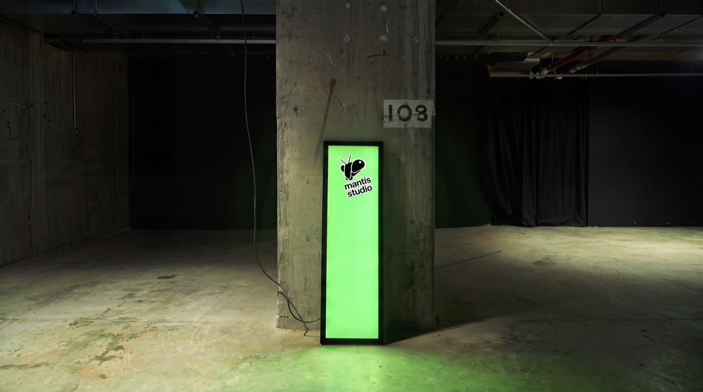 A tall green lightbox leaning against a concrete pillar numbered 108 in a dark, empty garage. The Mantis Studio logo sits at the top of its glowing face.